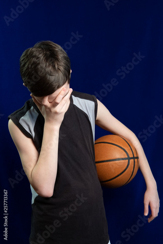 Basketball player. Ball in hand. Failure or frustration in a sports game. A teenager in a black T-shirt covered his face with his hand. Blue background.