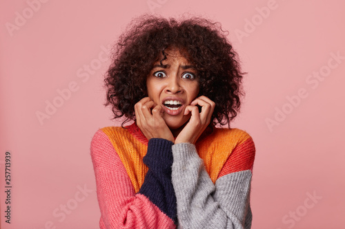 Close up portrait of scared hysterical african american girl with afro hairstyle looking frightened, in panic, nervous, scared, keeps fists near her face, isolated over pink background photo