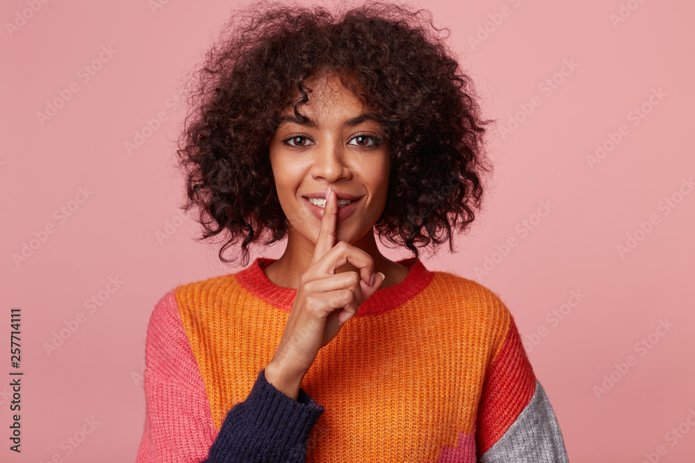Intriguing mysterious girl with a playful smile flirting, demonstrates a gesture of silence, holding an index finger near the mouth calls for privacy to be kept, secret, stay quiet,calm. Intriguing mysterious girl with a playful smile flirting, demonstrates a gesture of silence, holding an index finger near the mouth calls for privacy to be kept, secret, stay quiet,calm.