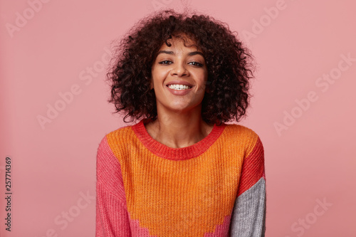 Positive optimistic charming african american girl looking swell wonderful playful confident joyful, with afro hairstyle, with lively smile, wearing colorful longsleeve, isolated on pink background