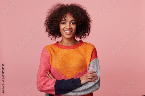 Studio photo of charismatic attractive african american girl with afro hairstyle looks with pleasure, with plesant smile, stand with arms crossed, wearing colorful longsleeve, isolated on pink wall photo