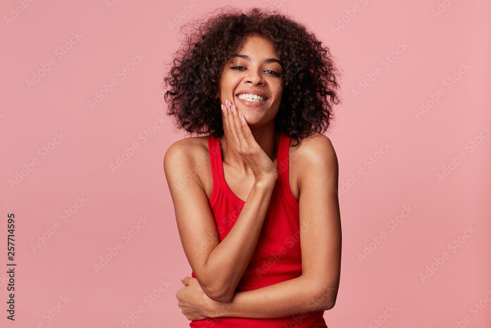 Happy pleased charming african american girl with afro hairstyle looks with pleasure, touches her face with palm, smiles, rejoices from soft skin, wearing red singlet, isolated on pink background