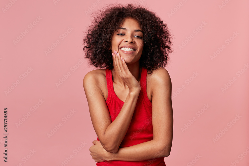 Happy pleased charming african american girl with afro hairstyle looks with pleasure, touches her face with palm, smiles, rejoices from soft skin, wearing red singlet, isolated on pink background Happy pleased charming african american girl with afro hairstyle looks with pleasure, touches her face with palm, smiles, rejoices from soft skin, wearing red singlet, isolated on pink background