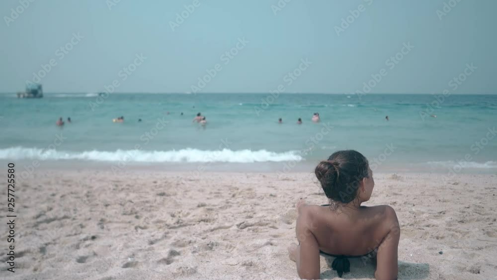brunette in black bikini with open shoulders lies on yellow sandy beach under clear blue sky backside view brunette in black bikini with open shoulders lies on yellow sandy beach under clear blue sky backside view