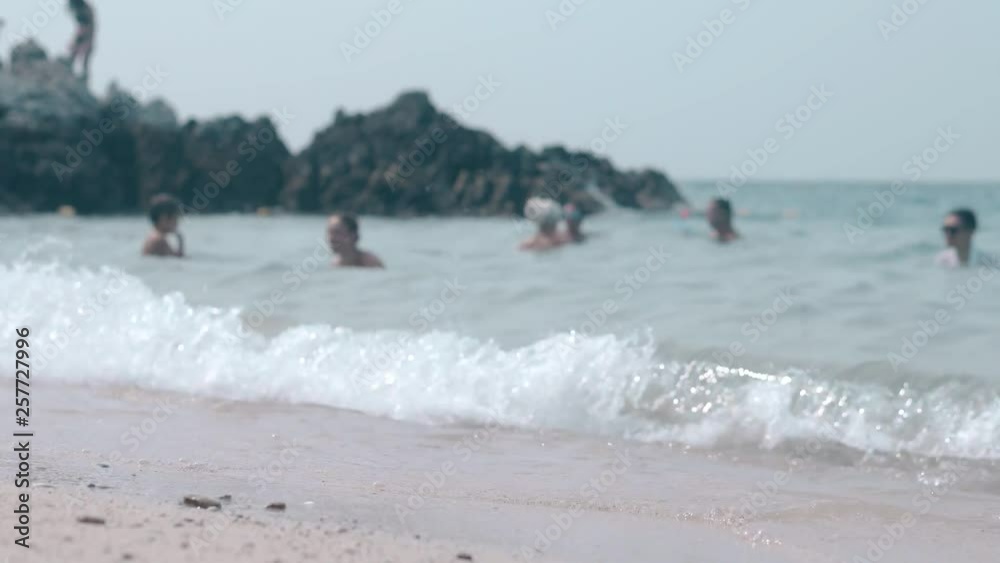 person in blue shorts walks on sandy beach against endless ocean with swimming people and brown rock person in blue shorts walks on sandy beach against endless ocean with swimming people and brown rock