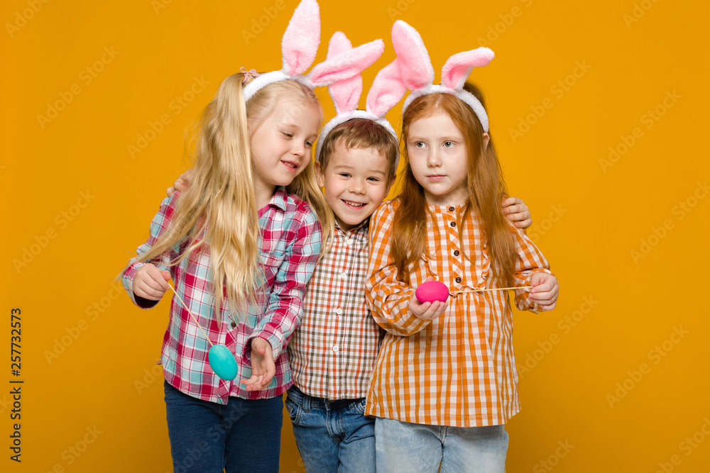 two little girls and boy with Easter bunny ears holding colorful eggs on yellow background. Happy easter. cute children two little girls and boy with Easter bunny ears holding colorful eggs on yellow background. Happy easter. cute children