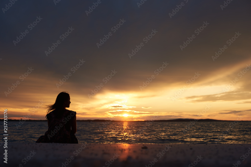 Back view silhouette of tourist woman sitting alone on seashore at water edge, enjoying beautiful view of sunset on dark evening sky background. Tourism and vacations concept.
