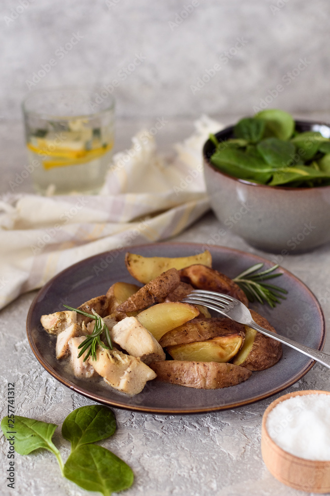 Baked potatoes sliced ​​with chicken fillet in a creamy sauce in a plate with a fork on the background of a salad bowl with spinach and a glass of water and lemon.