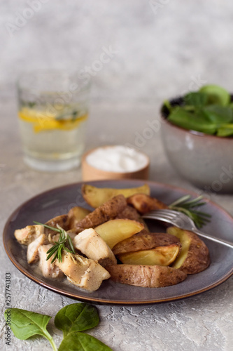 Baked potatoes sliced ​​with chicken fillet in a creamy sauce in a plate and salad bowl with spinach, a glass with water and lemon on a gray background.
