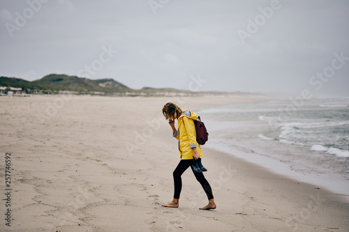 Young woman wearing yellow rain jacket speaking over the mobile phone while strolling along North Sea coast barefoot carrying her shoes in the hand