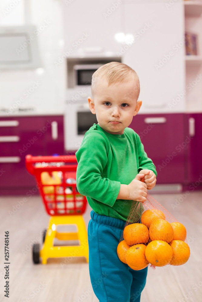 Little boy holds a grid with oranges. Little kid in casual wear carrying child plastic shopping trolley. Little boy holds a grid with oranges. Little kid in casual wear carrying child plastic shopping trolley.