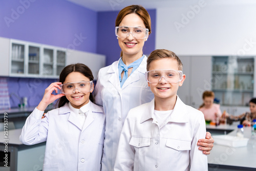 Smiling teacher in white coat and protective goggles embracing pupils during chemistry lesson