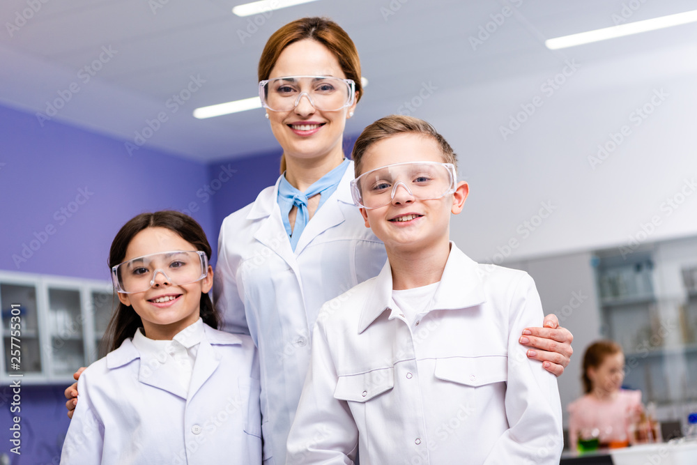 Smiling teacher in white coat and protective goggles embracing pupils during chemistry lesson Smiling teacher in white coat and protective goggles embracing pupils during chemistry lesson
