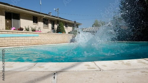 Young fit Caucasian man runs off diving board and jumps into a swimming pool in a backyard. Man in swimming trunks jumps into an outdoor swimming pool. Jumping into backyard pool making a splash.