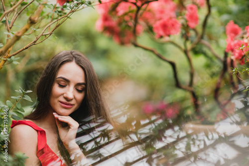 Young beautiful caucasian woman in glass greenhouse among colorful azalea flowers. Art portrait of a long-haired girl wearing a romantic red dress.