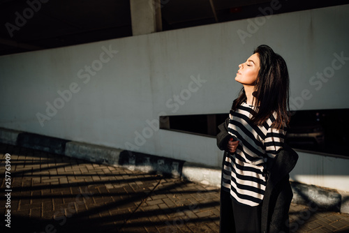 beautiful young happy girl in black pants and striped vest on parking lot, brunette woman is resting outdoors