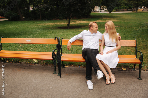 Beautiful couple sits on the bench in the park. Blonde hair man and woman spend time togerther. They take black umbrella. Background of green grass