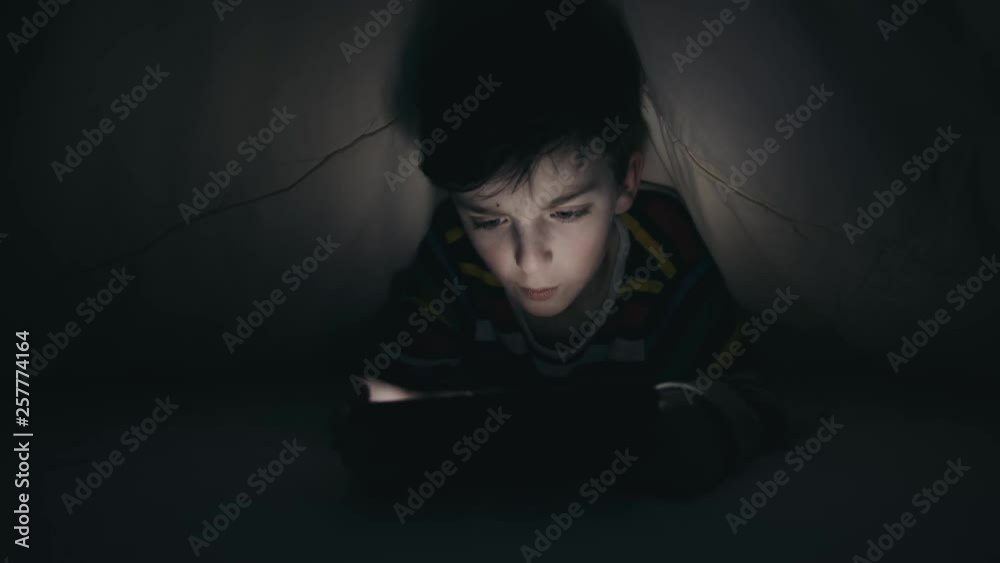 Happy boy playing emotionally game on smartphone in pajamas lying in bed under yellow blanket and on tablet in dark. Child plays at bedtime in the evening. Internet. Gamer. Hobby Happy boy playing emotionally game on smartphone in pajamas lying in bed under yellow blanket and on tablet in dark. Child plays at bedtime in the evening. Internet. Gamer. Hobby