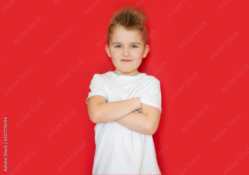 A little boy in a white shirt stands with his arms folded cross. Red background.
