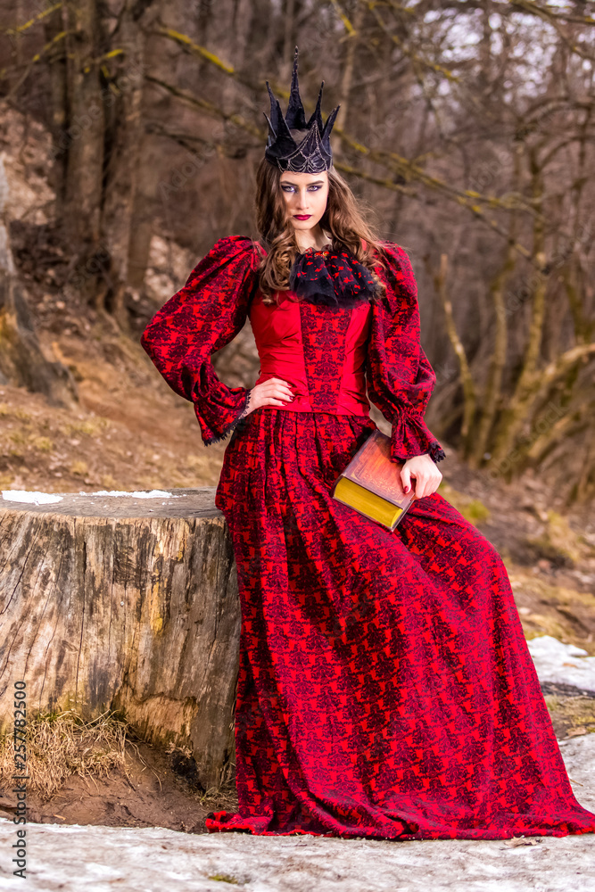 Art Photography. Mysterious Medieval Queen in Red Dress and Spiky Black Crown Posing With Ancient Book in Forest in Early Spring.
