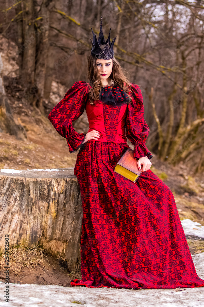 Art Photography. Mysterious Medieval Queen in Red Dress and Spiky Black Crown Posing With Ancient Book in Forest in Early Spring. Art Photography. Mysterious Medieval Queen in Red Dress and Spiky Black Crown Posing With Ancient Book in Forest in Early Spring.