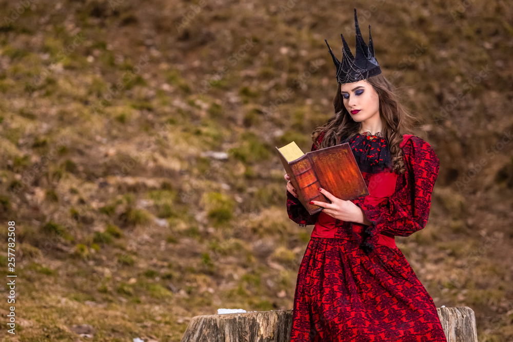 Art Photography. Mysterious Fairy Medieval Queen in Red Dress and Spiky Black Crown Posing With Ancient Book in Forest in Early Spring.