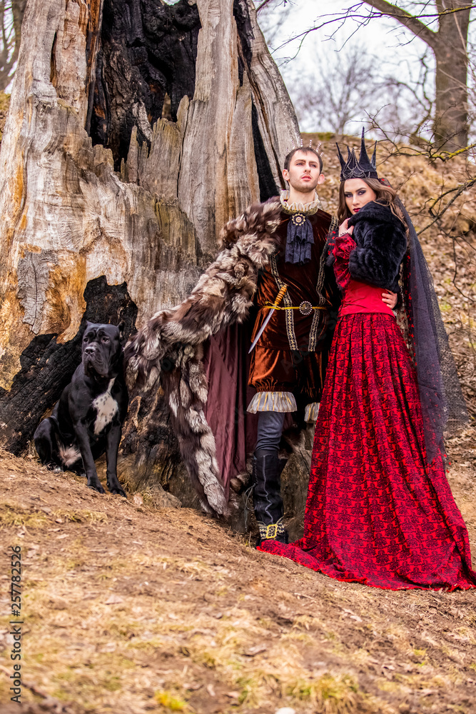 Costume Play Project. Caucasian Couple as Prince and Princess with Black Dog Posing in Medieval Clothing in Spring Forest Outdoors Costume Play Project. Caucasian Couple as Prince and Princess with Black Dog Posing in Medieval Clothing in Spring Forest Outdoors