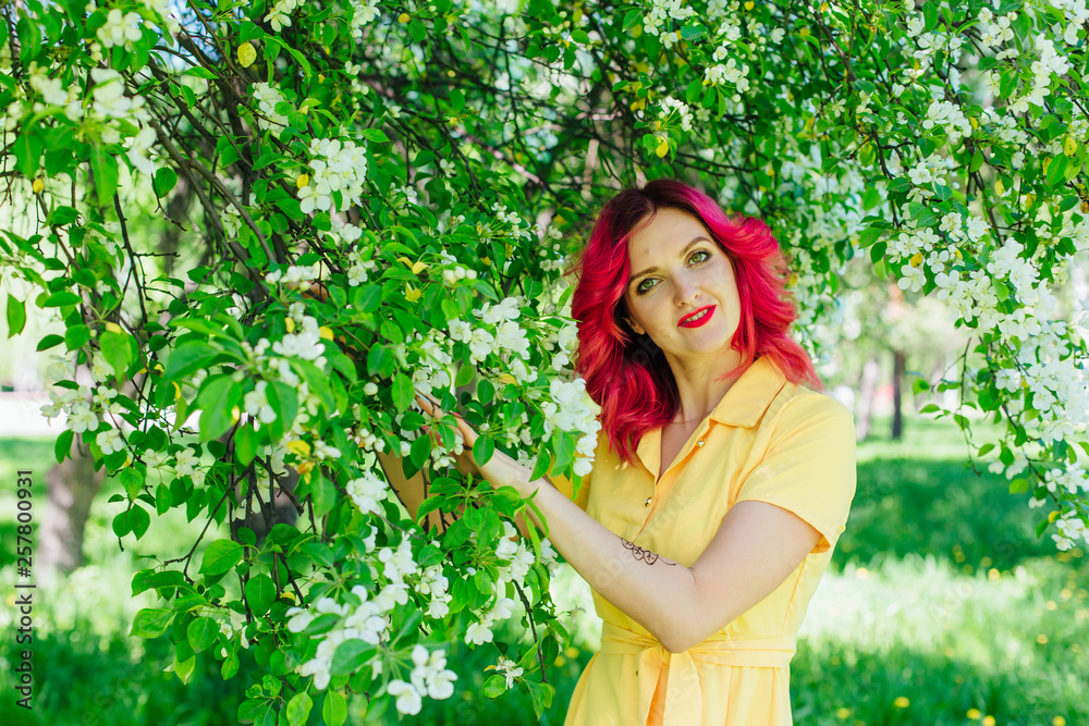 Beautiful and young woman with bright red hair and ref lips standing next to a blooming apple tree in a yellow dress. Beautiful and young woman with bright red hair and ref lips standing next to a blooming apple tree in a yellow dress.
