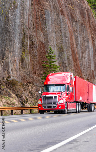 Bright red big rig semi truck with semi trailer running on winding mountain road with high cliff on the side