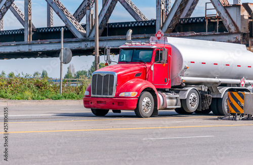 Red local big rig semi truck with day cab transports fuel in the tank trailer running under the old bridge