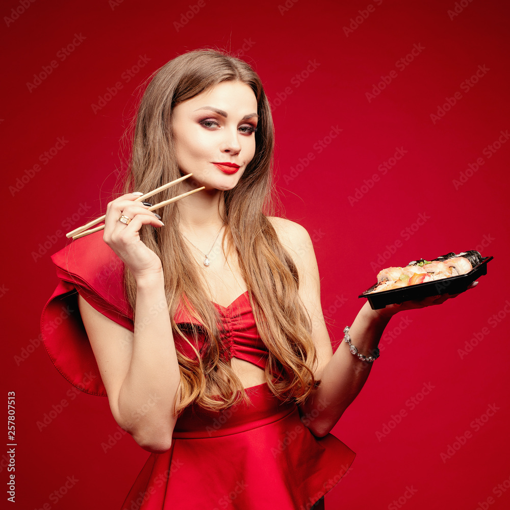 Front view of gorgeous woman in sexy red dress eating tasty sushi on red  isolated background. Young female enjoying eastern food, looking at camera  and smiling in studio. Concept of enjoyment. Stock