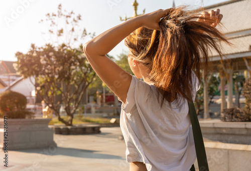 Behind view of an Asian girl student tying her hair walking through a urban college campus - Female ethnic Thai woman bunching her hair into a ponytail walking in a park in bright summer sunshine 