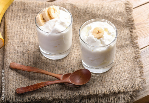 Two glasses of white yogurt with banana slices and wooden spoons on rough burlap napkin. 