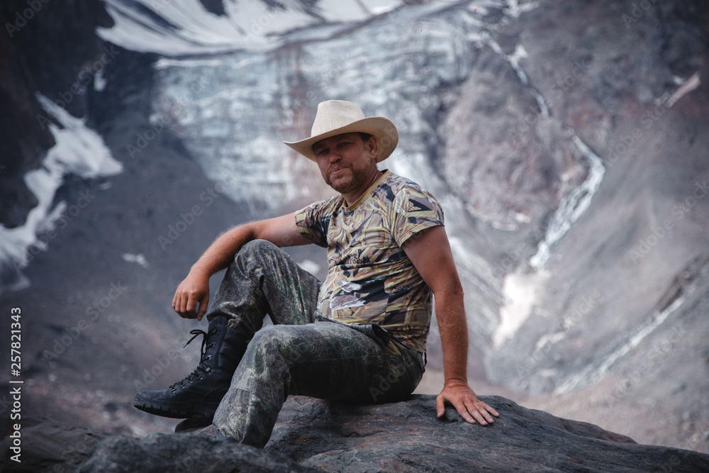 a middle-aged man in a cowboy hat sits on a rock against a glacier and a mountain