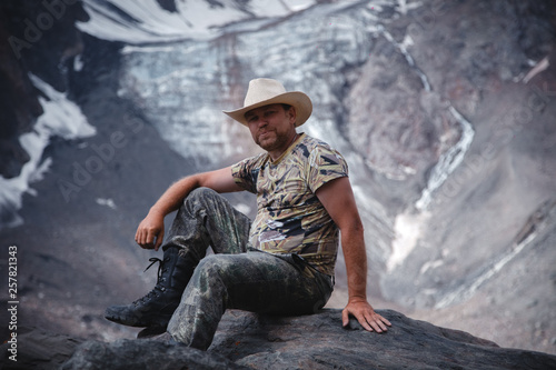 a middle-aged man in a cowboy hat sits on a rock against a glacier and a mountain