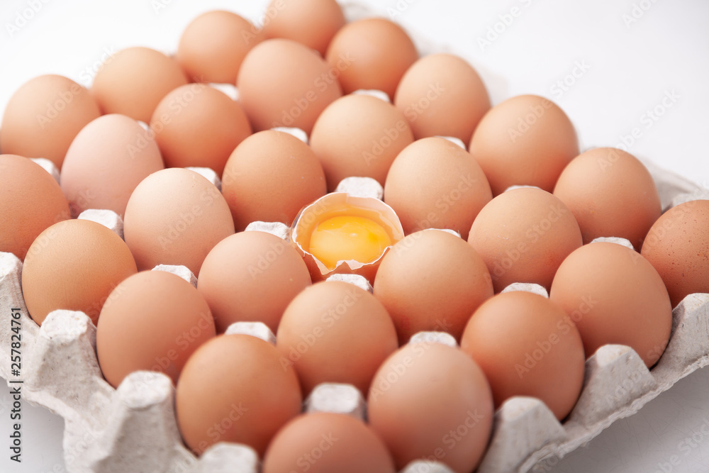  A tray of whole chicken eggs and one broken on a white background.