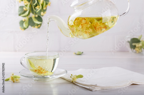 Pouring linden tea from teapot into transparent cup. Glass cup and tea pot with herbal lime tree tea on white background. Medicinal plant, flowers used for herbal teas and tinctures.