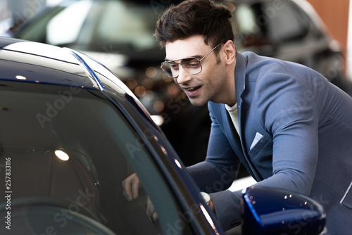 happy customer in glasses standing near automobile in car showroom ac © LIGHTFIELD STUDIOS