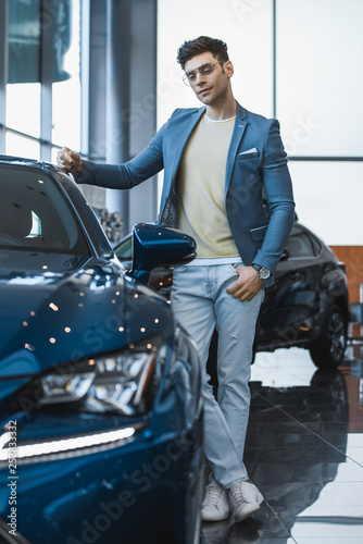 selective focus of man in glasses standing near automobile with crossed legs in car showroom