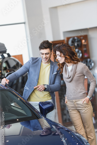 happy stylish man in glasses pointing with finger at automobile near attractive woman with hands in pockets