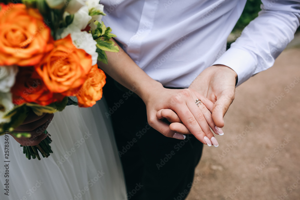 Wedding couple in love is holding a bouquet. Bride's hand with french manicure and engagement ring. Groom hugging his wife. Young in love. Summer day outdoors. Wedding couple in love is holding a bouquet. Bride's hand with french manicure and engagement ring. Groom hugging his wife. Young in love. Summer day outdoors.