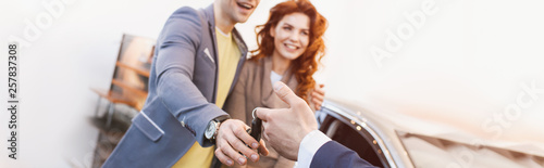 panoramic shot of car dealer giving keys to cheerful man and woman in car showroom
