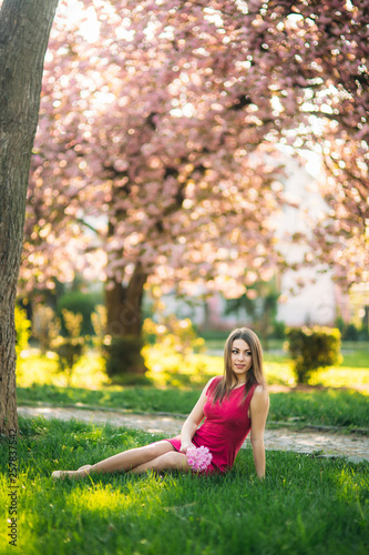 Young girl sitting on the grass and hold a sakura flowers in her hands. Background of pink bokeh of sakura trees