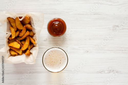 Fried potato wedges in paper box, barbecue sauce and glass of cold beer on a white wooden background, top view. Flat lay, overhead. Copy space.