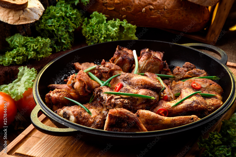Roasted chicken legs in a black cauldron on a wooden board among fresh vegetables on a dark wooden background. Vegetable composition. Chicken BBQ. Close-up.