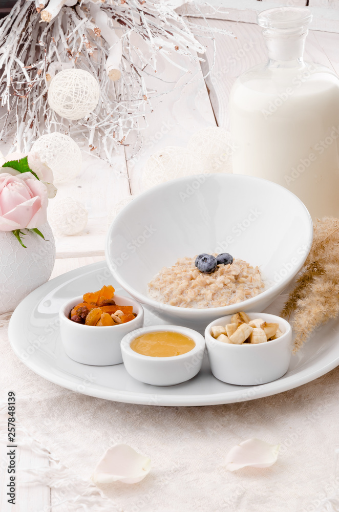 Oatmeal with blueberries in a beautiful white plate with honey, berries and a banana on a light delicate background, decorated with live roses. Beautiful healthy breakfast. Close-up. Space Oatmeal with blueberries in a beautiful white plate with honey, berries and a banana on a light delicate background, decorated with live roses. Beautiful healthy breakfast. Close-up. Space