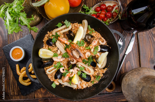 Traditional Spanish dish - Paella with seafood: shrimps, squids, mussels; wine, lemon and rice in a black cauldron on a dark rustic wooden background. Close up. Space. 