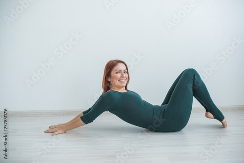 Beautiful flexible woman doing yoga exercises on white background. Red-haired woman in a green jumpsuit engaged in gymnastics