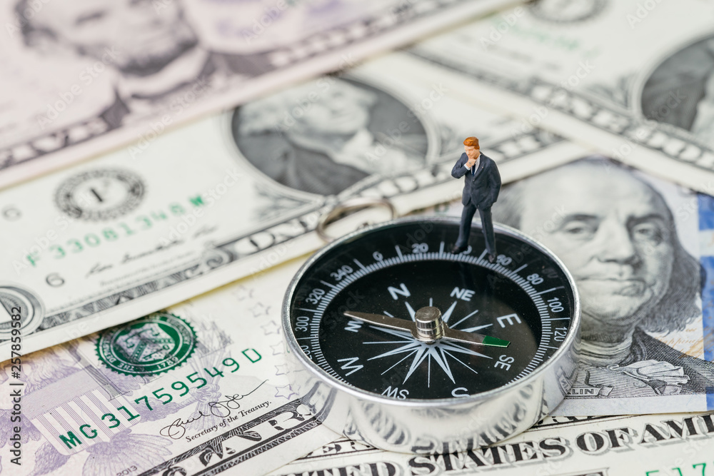 Miniature businessman in suit standing and thinking about future on compass and pile of US dollar money banknote, investment, financial crisis or economic direction concept Miniature businessman in suit standing and thinking about future on compass and pile of US dollar money banknote, investment, financial crisis or economic direction concept