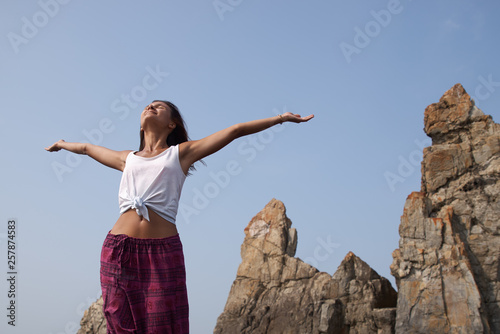 A slender young woman catches the sea breeze with her hands and enjoys the warm wind against the blue clear sky. The concept of freedom, outdoor recreation among the mountains and rocks